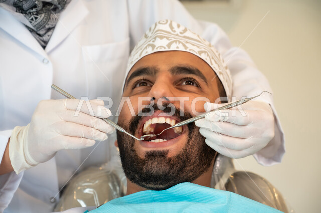 تقديم الخدمات والرعاية الصحية في المراكز التخصصية، الحصول على ابتسامة مثالية, شاب عربي خليجي عماني يرتدي الكمه يجلس في العيادة السنية ويفتح فمه لتشخيص المرض, ايدي طبيبة ترتدي القفازات الطبية تقوم بفحص أسنان المريض باستخدام الأدوات اللازمة