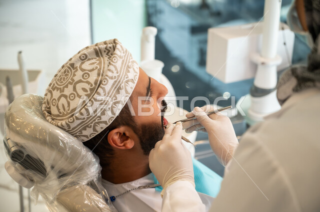 تقديم الخدمات والرعاية الصحية في المراكز التخصصية، الحصول على ابتسامة مثالية, شاب عربي خليجي عماني يرتدي الكمه يجلس في العيادة السنية ويفتح فمه لتشخيص المرض, صورة مقربة من الخلف لطبيبة ترتدي القفازات الطبية تقوم بفحص أسنان المريض باستخدام الأدوات اللازمة