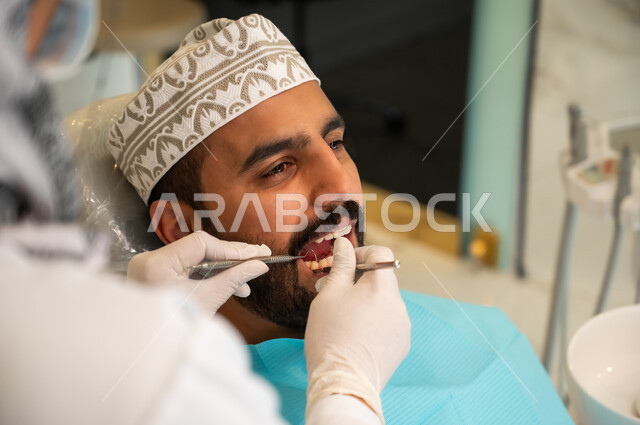 تقديم الخدمات والرعاية الصحية في المراكز التخصصية، الحصول على ابتسامة مثالية, شاب عربي خليجي عماني يرتدي الكمه يجلس في العيادة السنية ويفتح فمه لتشخيص المرض, صورة مقربة من الخلف لطبيبة ترتدي القفازات الطبية تقوم بفحص أسنان المريض باستخدام الأدوات اللازمة