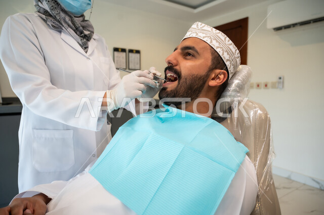 Equipment and tools needed for examinations, treatment of caries and decay, close-up of a young Omani Gulf Arab man wearing traditional clothing sitting in a dental clinic and opening his mouth to diagnose the disease, a female Omani Gulf Arab doctor wearing medical gloves examines the patient's teeth