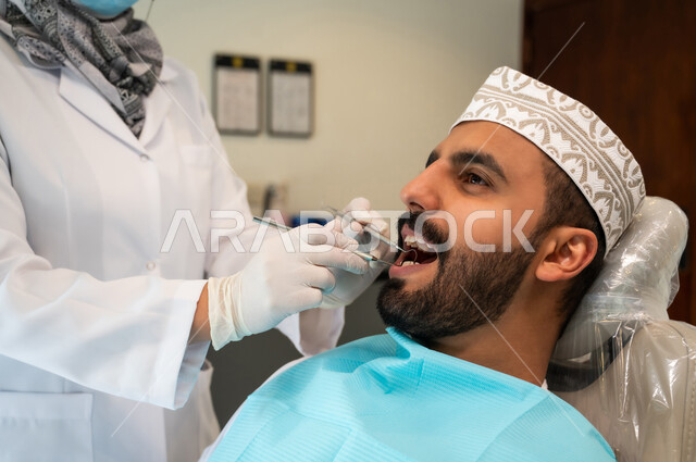 معالجة النخر والتسوس, صورة مقربة لشاب عربي خليجي عماني يرتدي الزي التقليدي يجلس في العيادة السنية ويفتح فمه لتشخيص المرض, طبيبة عربية خليجيه عمانية ترتدي القفازات الطبية تقوم بفحص أسنان المريض, الادوات والمعدات اللازمة لإجراء الفحوصات