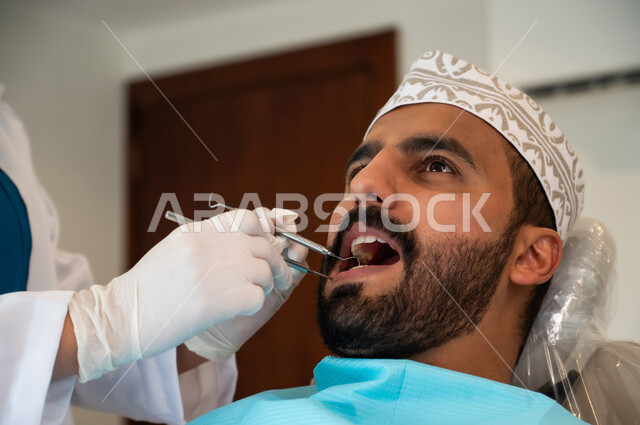 الحصول على ابتسامة مثالية، الادوات اللازمة لإجراء الفحوصات، معالجة النخر والتسوس, صورة مقربة لشاب عربي خليجي عماني يرتدي الزي التقليدي يجلس في العيادة السنية ويفتح فمه لتشخيص المرض, ايدي طبيبة عربية خليجيه عمانية ترتدي القفازات الطبية تقوم بفحص أسنان المريض