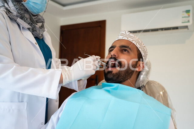 معالجة النخر والتسوس, صورة مقربة لشاب عربي خليجي عماني يرتدي الزي التقليدي يجلس في العيادة السنية ويفتح فمه لتشخيص المرض, طبيبة عربية خليجيه عمانية ترتدي القفازات الطبية تقوم بفحص أسنان المريض, الادوات والمعدات اللازمة لإجراء الفحوصات