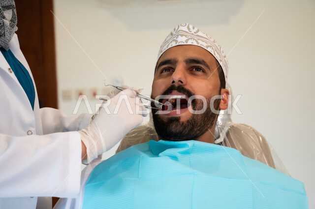 الحصول على ابتسامة مثالية، الادوات اللازمة لإجراء الفحوصات، معالجة النخر والتسوس, صورة مقربة لشاب عربي خليجي عماني يرتدي الزي التقليدي يجلس في العيادة السنية ويفتح فمه لتشخيص المرض, ايدي طبيبة عربية خليجيه عمانية ترتدي القفازات الطبية تقوم بفحص أسنان المريض