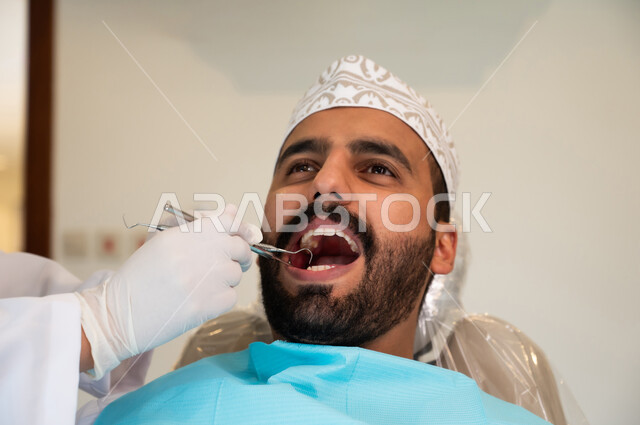 تقديم الخدمات والرعاية الصحية في المراكز التخصصية، الأساليب العلاجية المتبعة للعناية بالفم، الاهتمام والعناية بالصحة الفموية، استخدام الأدوات المعقمة لفحص أسنان المريض، صورة مقربة لشاب عربي خليجي عماني يرتدي الزي التقليدي يجلس على كرسي العلاج