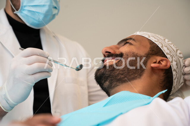 Using sterile instruments for dental examinations, oral health care, concept of medicine and healthcare, providing medical services to patients, doctor wearing gloves and a mask examining a patient's teeth, close-up of a young Omani Gulf Arab man wearing traditional clothing sitting on a treatment chair