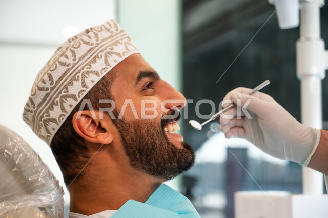 ايماءات البشاشة والسرور، تقديم الخدمات والرعاية الصحية في المراكز التخصصية، الأساليب العلاجية المتبعة للعناية بالفم، استخدام الأدوات المعقمة لفحص أسنان المريض، صورة مقربة من الجانب لشاب عربي خليجي عماني يرتدي الزي التقليدي يجلس على كرسي العلاج