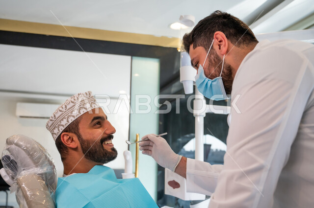 Conducting routine oral health checkups, providing medical services to patients, a doctor wearing gloves and a mask examining a patient's teeth, a close-up side view of a young Omani Gulf Arab man wearing a dishdasha and a kuma sitting on a dental chair.