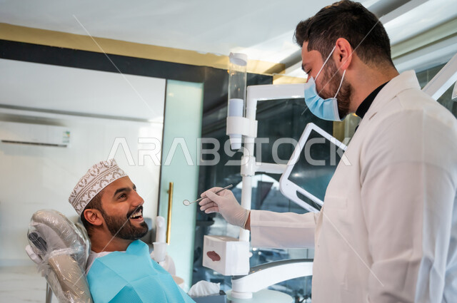 إجراء الفحوصات الدورية للعناية بالصحة الفموية، تقديم الخدمات الطبية للمرضى,  طبيب يرتدي القفازات والكمامة يقوم بفحص أسنان المريض، صورة مقربة من الجانب لشاب عربي خليجي عماني يرتدي الدشداشة والكمة يجلس على كرسي العلاج في العيادة السنية.