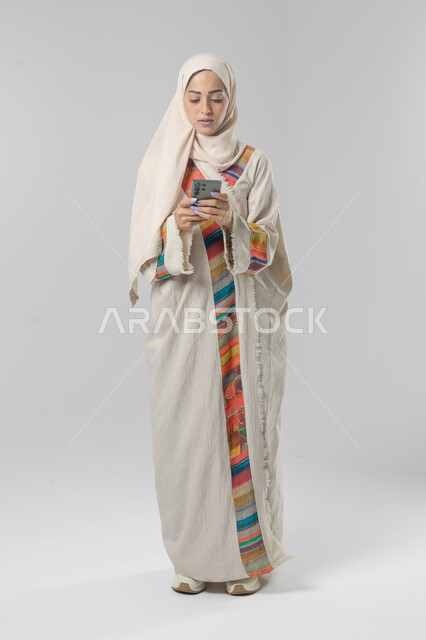  تعابير وايماءات الاندماج والتركيز في الجوال، استخدام التقنية الحديثه المتطورة في الحياة العملية، بورتريه لإمرأة عربية خليجية سعودية ترتدي الحجاب والعباءة تقوم بالعمل على الهاتف المحمول، صورة الجسم بالطول الكامل، خلفية بيضاء 