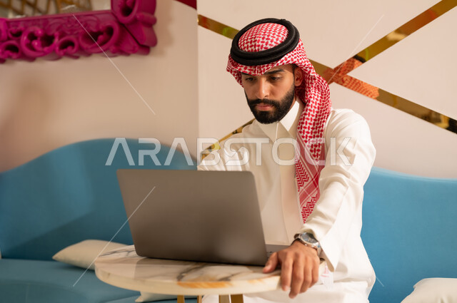 Expressions of integration and concentration in completing work tasks via laptop, calm atmosphere outside the home, cafes in the Kingdom of Saudi Arabia, a Saudi Arabian Gulf Arab man wearing a thobe and shemagh sitting on the couch browsing the internet using the laptop