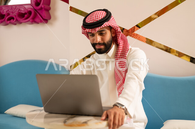 الاستمتاع بالاجواء الهادئه خارج المنزل، مقاهي في المملكة العربية السعودية، رجل عربي خليجي سعودي يرتدي الثوب و الشماغ يجلس على الأريكة يقوم بإنجاز مهام العمل عبر جهاز اللابتوب بإيماءات الاندماج و التركيز، تصفح الانترنت باستخدام الحاسوب المحمول
