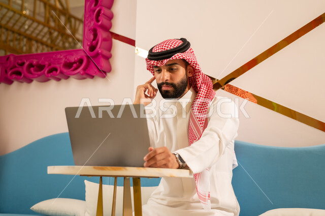 ايماءات الاندماج و التركيز والتفكير، انجاز مهام العمل عبر جهاز اللابتوب، اجواء هادئه خارج المنزل، مقاهي في المملكة العربية السعودية، رجل عربي خليجي سعودي يرتدي الثوب و الشماغ يجلس على الأريكة يقوم بتصفح الانترنت باستخدام الحاسوب المحمول