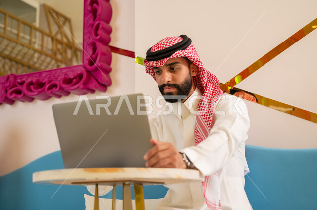 تصفح مواقع الانترنت باستخدام الحاسوب المحمول، الاستمتاع بالاجواء الهادئه خارج المنزل، مقاهي في المملكة العربية السعودية، رجل عربي خليجي سعودي يرتدي الثوب و الشماغ يجلس على الأريكة يقوم بإنجاز مهام العمل عبر جهاز اللابتوب بإيماءات الاندماج و التركيز