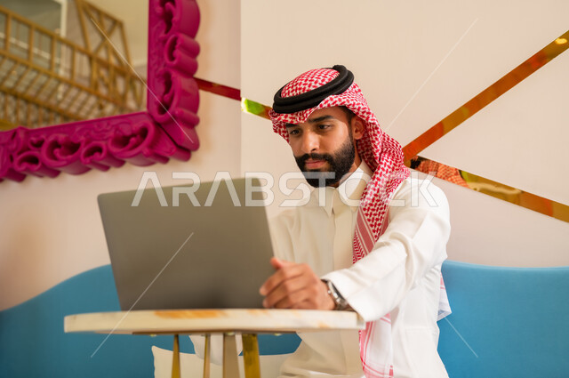 تعابير الاندماج و التركيز في انجاز مهام العمل عبر جهاز اللابتوب، اجواء هادئه خارج المنزل، مقاهي في المملكة العربية السعودية، رجل عربي خليجي سعودي يرتدي الثوب و الشماغ يجلس على الأريكة يقوم بتصفح الانترنت باستخدام الحاسوب المحمول