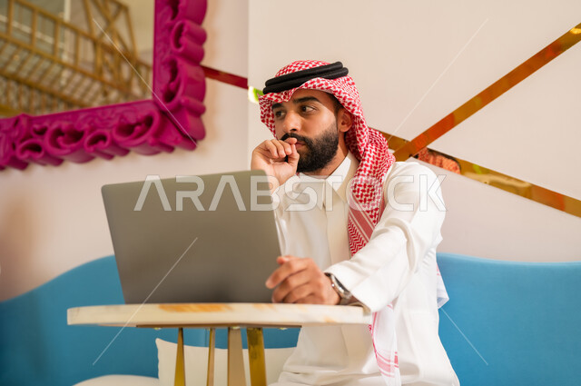 Looking at something with gestures of integration, concentration and thinking, completing work tasks via laptop, quiet atmosphere outside the home, cafes in Saudi Arabia, a Saudi Arabian Gulf Arab man wearing a thobe and shemagh sitting on the couch browsing the internet using the laptop