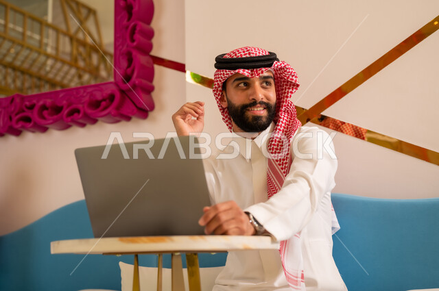 استخدام الحاسوب المحمول لإنجاز المشاريع في المقهى، الاستمتاع بالاجواء الهادئه خارج المنزل، مقاهي في المملكة العربية السعودية، رجل عربي خليجي سعودي يرتدي الثوب والشماغ يجلس على الأريكة ينظر إلى شيء ما بإيماءات السرور