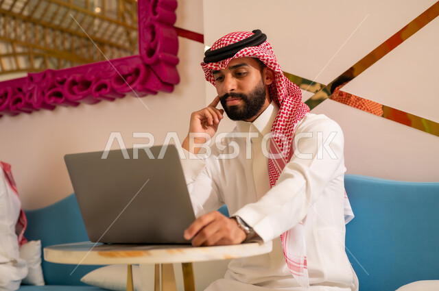 ايماءات الاندماج و التركيز والتفكير، انجاز مهام العمل عبر جهاز اللابتوب، اجواء هادئه خارج المنزل، مقاهي في المملكة العربية السعودية، رجل عربي خليجي سعودي يرتدي الثوب و الشماغ يجلس على الأريكة يقوم بتصفح الانترنت باستخدام الحاسوب المحمول