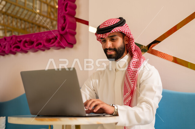 مطاعم ومقاهي في المملكة العربية السعودية، استخدام الحاسوب المحمول لإنجاز الأعمال والمشاريع، الاستمتاع بالاجواء الهادئه خارج المنزل، رجل عربي خليجي سعودي يرتدي الثوب والشماغ يجلس على الأريكة يقووم بتصفح مواقع التواصل عبر الانترنت بإيماءات الاندماج والتركيز