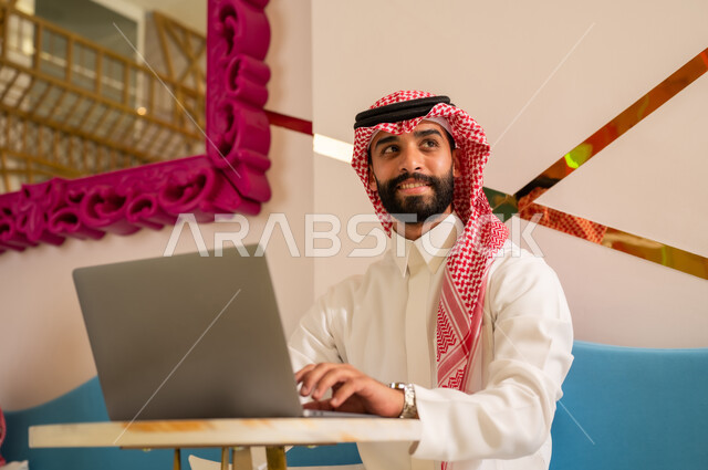 استخدام الحاسوب المحمول لإنجاز المشاريع في المقهى، الاستمتاع بالاجواء الهادئه خارج المنزل، مقاهي في المملكة العربية السعودية، رجل عربي خليجي سعودي يرتدي الثوب والشماغ يجلس على الأريكة ينظر إلى شيء ما بإيماءات السرور