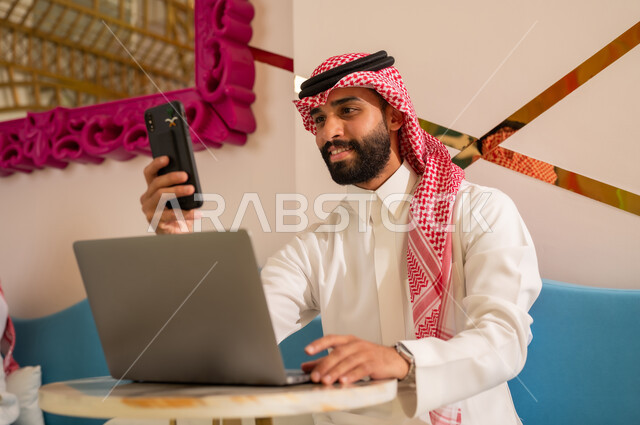 إدارة شؤون الأعمال وتنظيمها عبر الأجهزة التقنية الحديثة، تصفح مواقع التواصل عبر الانترنت بإيماءات السرور، إنجاز مهام العمل عن بعد، رجل عربي خليجي سعودي يرتدي الثوب والشماغ يجلس على الأريكة في إحدى المقاهي الراقية ويستخدم الهاتف والحاسوب المحمول