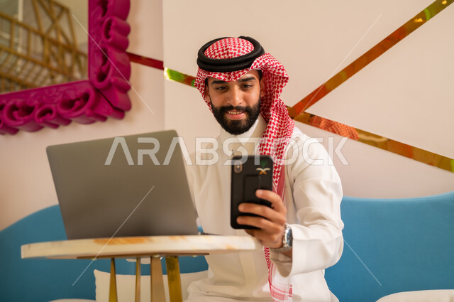 تصفح مواقع التواصل عبر الانترنت بإيماءات السرور، إنجاز مهام العمل عن بعد، إدارة شؤون الأعمال وتنظيمها عبر الأجهزة التقنية الحديثة، رجل عربي خليجي سعودي يرتدي الثوب والشماغ يجلس على الأريكة في إحدى المقاهي الراقية ويستخدم الهاتف والحاسوب المحمول