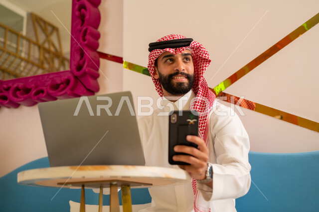 النظر إلى شيء ما بتعابير السرور، إدارة شؤون الأعمال وتنظيمها عبر الأجهزة التقنية الحديثة، تصفح مواقع التواصل عبر الانترنت، إنجاز مهام العمل عن بعد، رجل عربي خليجي سعودي يرتدي الثوب والشماغ يجلس على الأريكة في إحدى المقاهي الراقية ويستخدم الهاتف والحاسوب المحمول