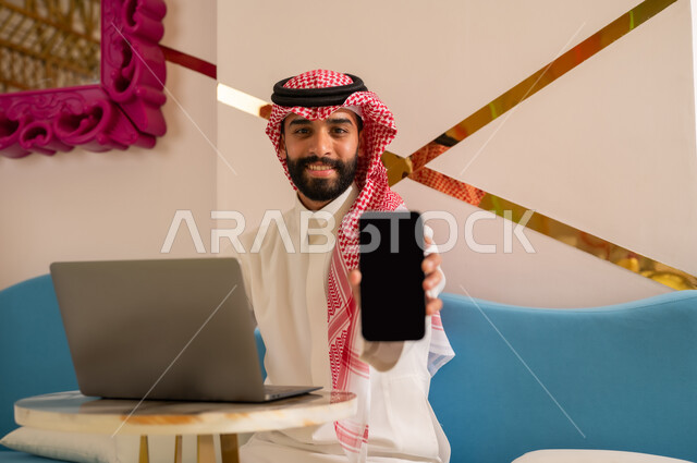 النظر إلى الكاميرا بإيماءات السرور والسعادة، الإعجاب بتطبيقات الأجهزة التقنية الحديثة، إنجاز مهام العمل عن بعد، رجل عربي خليجي سعودي يرتدي الثوب والشماغ يجلس على الأريكة في إحدى المقاهي الراقية يقوم بعرض شاشة سوداء فارغة عبر الهاتف المحمول