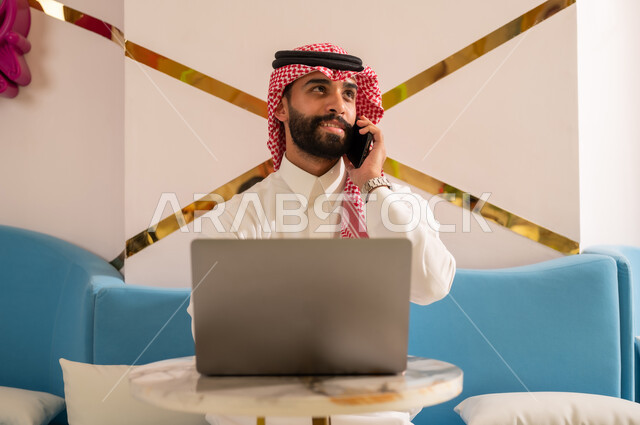 التواصل مع الأهل والأصدقاء، استخدام الأجهزة التقنية الحديثة، إدارة شؤون العمل عن بعد، رجل عربي خليجي سعودي مبتسم يرتدي الثوب والشماغ يجلس على الأريكة في إحدى المقاهي الراقية يقوم بإجراء مكالمة هاتفية عبر الهاتف المحمول
