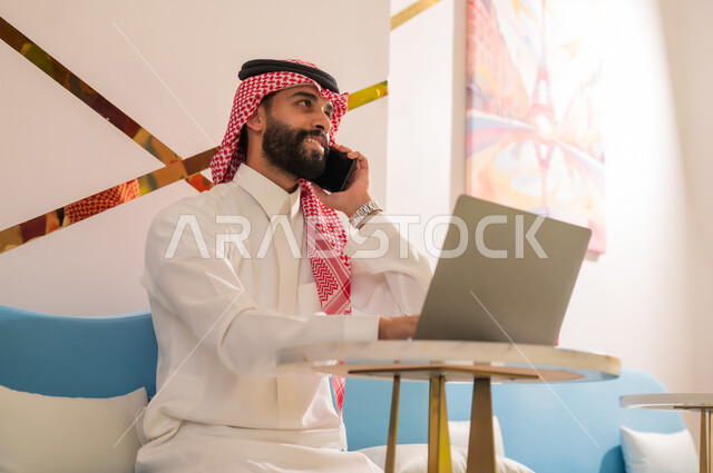 إدارة شؤون العمل عن بعد، التواصل مع الأهل والأصدقاء باستخدام الأجهزة التقنية الحديثة، رجل عربي خليجي سعودي مبتسم يرتدي الثوب والشماغ يجلس على الأريكة في إحدى المقاهي الراقية يقوم بإجراء مكالمة هاتفية عبر الهاتف المحمول
