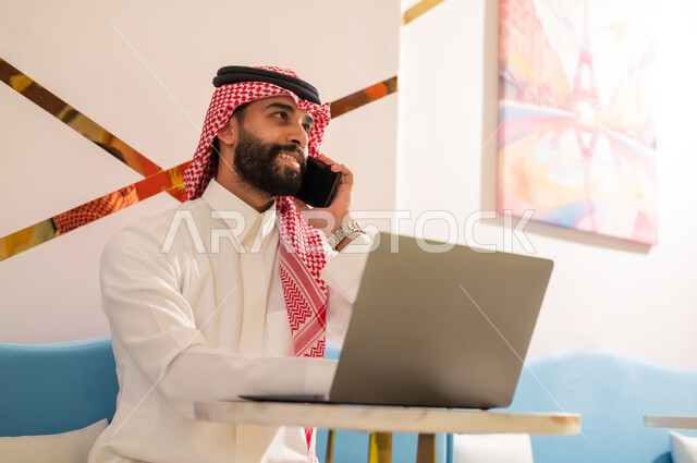 التواصل مع الأهل والأصدقاء، استخدام الأجهزة التقنية الحديثة، إدارة شؤون العمل عن بعد، رجل عربي خليجي سعودي مبتسم يرتدي الثوب والشماغ يجلس على الأريكة في إحدى المقاهي الراقية يقوم بإجراء مكالمة هاتفية عبر الهاتف المحمول