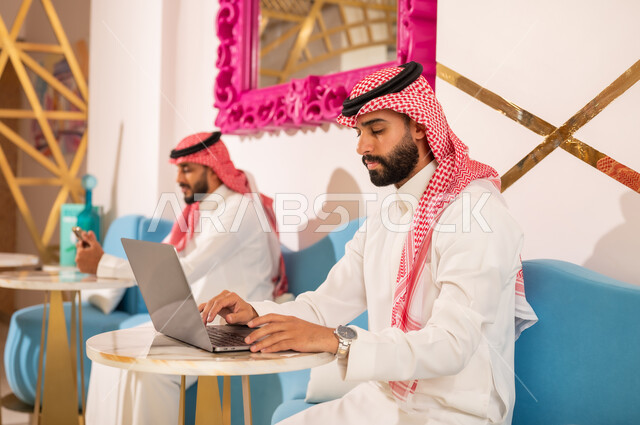 إيماءات الجدية والتركيز في العمل عبر الحاسوب المحمول، الوسائل التقنية الحديثة والمتطورة، شابان عربيان خليجيان سعوديان يرتديان الثوب والشماغ يجلسان في مقهى لإنجاز المهام عن بعد، قضاء وقت ممتع في الخارج، مطعم وكافيه راقي في المملكة العربية السعودية