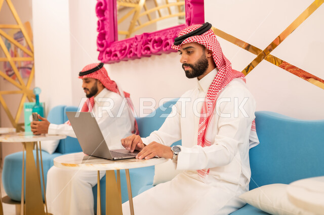 Gestures of seriousness and focus while working on a laptop, modern and advanced technical means, two young Saudi Arabian Gulf men wearing thobes and shemaghs sitting in a cafe to complete tasks remotely, spending a pleasant time outside, an upscale restaurant and cafe in the Kingdom of Saudi Arabia
