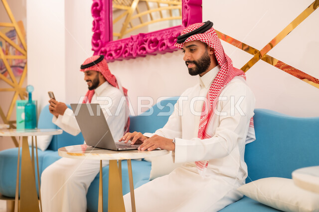 العمل عبر الانترنت باستخدام الحاسوب المحمول، الوسائل التقنية الحديثة والمتطورة، شابان عربيان خليجيان سعوديان يرتديان الثوب والشماغ يجلسان في مقهى لإنجاز المهام عن بعد، قضاء وقت ممتع في الخارج، مطعم وكافيه راقي في المملكة العربية السعودية