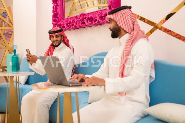 صداقة وتعارف وبناء علاقات اجتماعية، العمل عبر الانترنت باستخدام الحاسوب المحمول، الوسائل التقنية الحديثة والمتطورة، شابان عربيان خليجيان سعوديان يرتديان الثوب والشماغ يجلسان في مقهى لإنجاز المهام عن بعد، قضاء وقت ممتع في الخارج، مطعم وكافيه راقي في السعودية