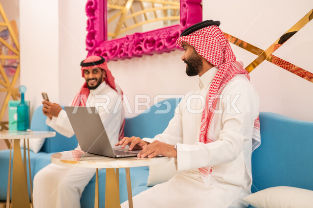 العمل عبر الانترنت باستخدام الحاسوب المحمول، الوسائل التقنية الحديثة والمتطورة، شابان عربيان خليجيان سعوديان يرتديان الثوب والشماغ يجلسان في مقهى لإنجاز المهام عن بعد، قضاء وقت ممتع في الخارج، مطعم وكافيه راقي في المملكة العربية السعودية