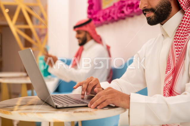 Upscale restaurant and cafe in Saudi Arabia, working on laptop, modern and advanced technology, close-up of two young Saudi Arabian Gulf men wearing thobes and shemaghs sitting in a cafe to complete tasks remotely, having a good time outside