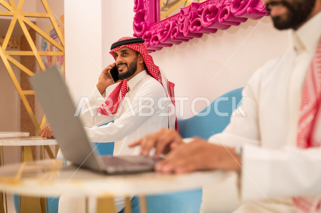 وسيلة تقنية حديثة ومتطورة، مطعم وكافيه راقي في المملكة العربية السعودية، العمل عبر الحاسوب المحمول، صورة مقربة لشاب عربي خليجي سعودي يرتدي الثوب والشماغ يجلس في مقهى لإنجاز المهام عن بعد، رجل يجري مكالمة هاتفية