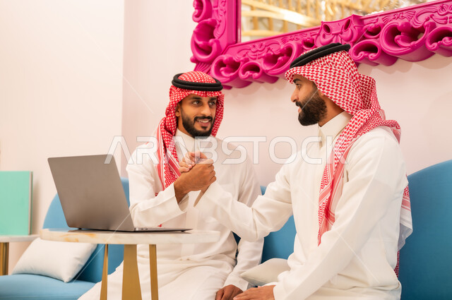 صداقة وتعارف وبناء علاقات اجتماعية، إنجاز مهام العمل عن بعد عبر الحاسوب المحمول، قضاء وقت ممتع في المقهى، عقد صفقات عمل جديدة ناجحة، رجلان عربيان خليجيان سعوديان يرتديان الزي التقليدي يقومان بمصافحة بعضهما بايماءات السعادة والسرور، استخدام جهاز تقني حديث ومتطور