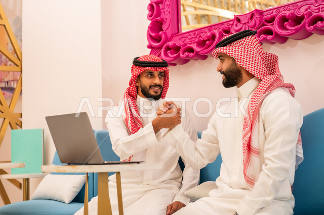 إنجاز مهام العمل عن بعد عبر الحاسوب المحمول، صداقات وتعارف وبناء علاقات اجتماعية، قضاء وقت ممتع في المقهى، عقد صفقات عمل جديدة ناجحة، رجلان عربيان خليجيان سعوديان يرتديان الزي التقليدي يقومان بمصافحة بعضهما بايماءات السعادة والسرور، استخدام جهاز تقني حديث ومتطور