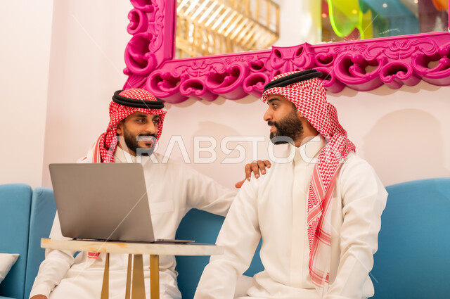 تبادل أطراف الحديث، صداقة وتعارف وبناء علاقات اجتماعية، العمل عبر الانترنت باستخدام الحاسوب المحمول، ايماءات المحبة والسرور، شابان عربيان خليجيان سعوديان يرتديان الثوب والشماغ يجلسان في مقهى لإنجاز المهام عن بعد، قضاء وقت ممتع في الخارج