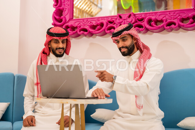 الإشارة إلى رؤية شيء ما على اللابتوب، انجاز المشاريع والمهام في الكافيه باستخدام جهاز الحاسوب المحمول، قضاء وقت ممتع مع الأصدقاء, الحوار والنقاش بين زملاء العمل، إيماءات السرور والسعادة، شاب عربي خليجي سعودي يرتدي الثوب والشماغ يجلس مع صديقه في المقهى.