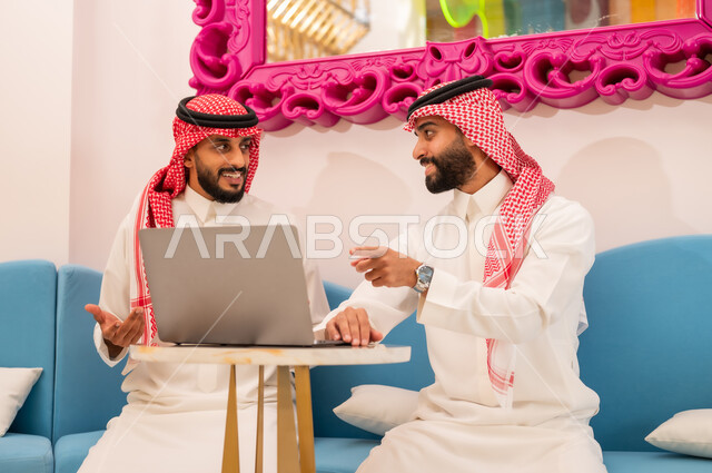 انجاز المشاريع والمهام في الكافيه باستخدام جهاز الحاسوب المحمول، الإشارة إلى رؤية شيء ما على اللابتوب، قضاء وقت ممتع مع الأصدقاء, الحوار والنقاش بين زملاء العمل، إيماءات السرور والسعادة، شاب عربي خليجي سعودي يرتدي الثوب والشماغ يجلس مع صديقه في المقهى.
