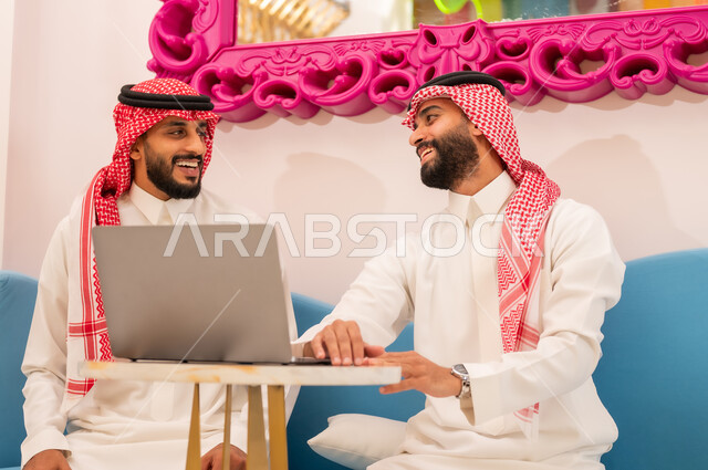انجاز المشاريع والمهام في الكافيه باستخدام جهاز الحاسوب المحمول، قضاء وقت ممتع مع الأصدقاء, الحوار والنقاش بين زملاء العمل، إيماءات السرور والسعادة، شاب عربي خليجي سعودي يرتدي الثوب والشماغ يجلس مع صديقه في المقهى.