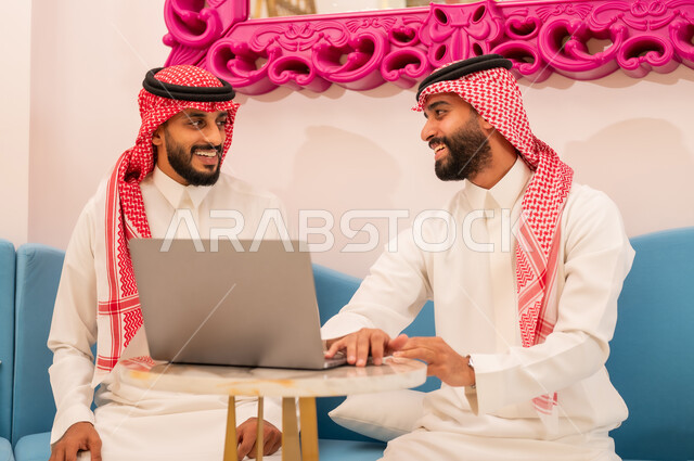 قضاء وقت ممتع مع الأصدقاء, الحوار والنقاش بين زملاء العمل، ايماءات السرور والسعاده، استخدام جهاز الحاسوب المحمول لانجاز المشاريع والمهام في الكافيه، شابان عربيان خليجيان سعوديان يرتديان الثوب والشماغ السعودي يجلسان على الأريكة في المقهى.