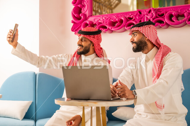 Completing work remotely using modern technical devices, documenting happy moments, spending quality time with friends, taking souvenir photos via mobile phone with gestures of pleasure, two Saudi Arabian Gulf friends wearing traditional dress sitting on the couch in an upscale cafe
