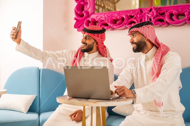 توثيق اللحظات السعيدة، قضاء وقت ممتع برفقة الأصدقاء، التقاط الصور التذكارية عبر الهاتف المحمول بإيماءات السرور، صديقان عربيان خليجيان سعوديان يرتديان الزي التقليدي يجلسان على الأريكة في إحدى المقاهي الراقية، إنجاز العمل عن بعد باستخدام الأجهزة التقنية الحديثة