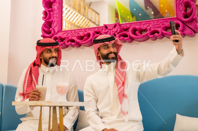 النظر إلى الكاميرا بإيماءات السرور والسعادة، قضاء وقت ممتع برفقة الأصدقاء خارج المنزل، مشاركة المشروبات الباردة، التقاط الصور التذكارية عبر الهاتف المحمول، صديقان عربيان خليجيان سعوديان يرتديان الزي التقليدي يجلسان على الأريكة في إحدى المقاهي الراقية