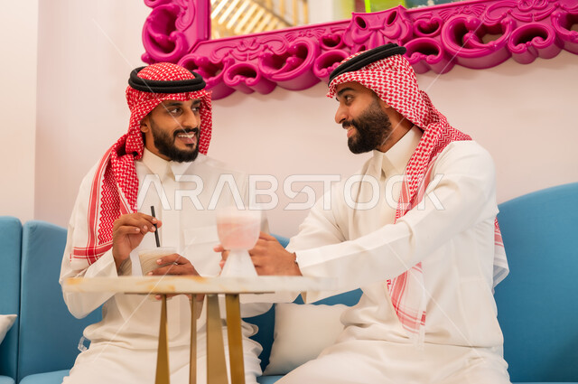 A relaxing weekend, friends meeting over cold drinks, gestures of happiness and pleasure, spending a good time with friends, two Saudi Arabian Gulf friends wearing traditional dress sitting on the couch in an upscale cafe, exchanging pleasantries