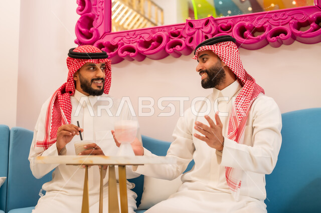 Friends meeting for cold drinks, a fun weekend session, gestures of happiness and pleasure, spending a good time with friends, two Saudi Arabian Gulf friends wearing traditional dress sitting on the couch in an upscale cafe exchanging pleasantries